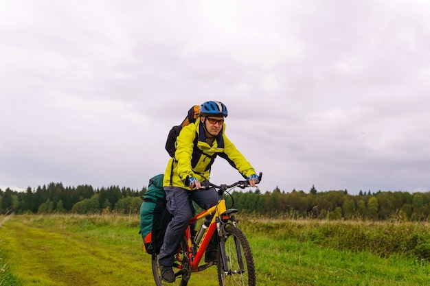 Bikepacker mâle monte sur un chemin de terre à travers un champ un jour d'automne nuageux