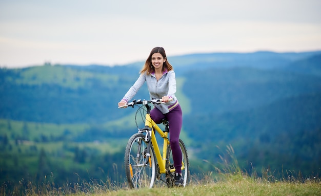 Biker femme forte à vélo sur un vélo de montagne jaune sur un sentier rural dans la soirée. Montagnes sur l'arrière-plan flou. Activité sportive en plein air, concept de style de vie