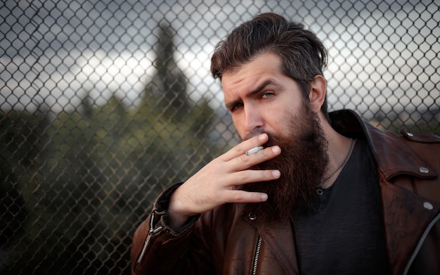 Biker masculin brutal avec une longue barbe, moustache et cheveux gris dans une veste en cuir marron fume une cigarette