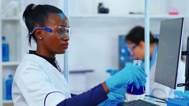 Biologiste de femme noire faisant des recherches à l'aide d'un ordinateur dans un laboratoire moderne équipé. Des trucs multiethniques examinant l'évolution des vaccins à l'aide de la haute technologie et de la recherche de traitements contre le virus covid19