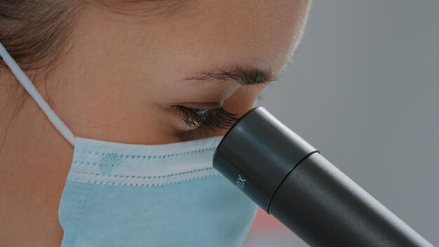 Biologiste avec masque facial utilisant une lentille microscopique sur un outil de chimie en laboratoire. Ingénieur scientifique utilisant un microscope avec une loupe et une mise au point sélective pour travailler sur une expérience en laboratoire.