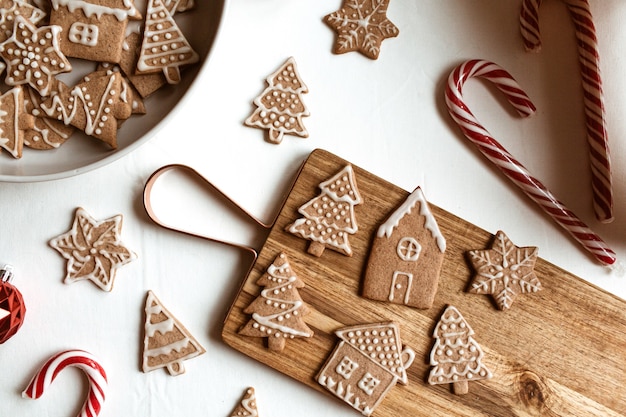 Biscuits au gingembre faits maison étoiles, sapins, maisons sur planche à découper en bois, bonbons bâton sur blanc. Mise à plat, composition de Noël vue de dessus.