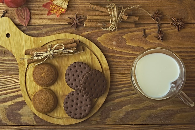 Biscuits aux pépites de chocolat sur un plateau et du lait dans une tasse de cuisson avec des bâtons de cannelle sur un fond en bois...