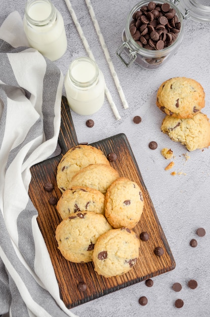 Biscuits maison traditionnels américains avec des pépites de chocolat sur une vieille planche de bois sur une surface grise avec des bouteilles de lait et de paille. Orientation verticale. Vue de dessus.