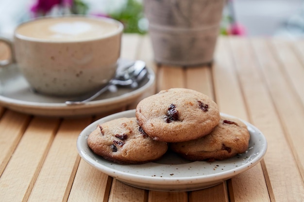 Biscuits naturels aux canneberges sans sucre sur l'assiette cappuccino dans une tasse en céramique blanche sur une table en bois à la terrasse du café Petit-déjeuner esthétique à l'extérieur