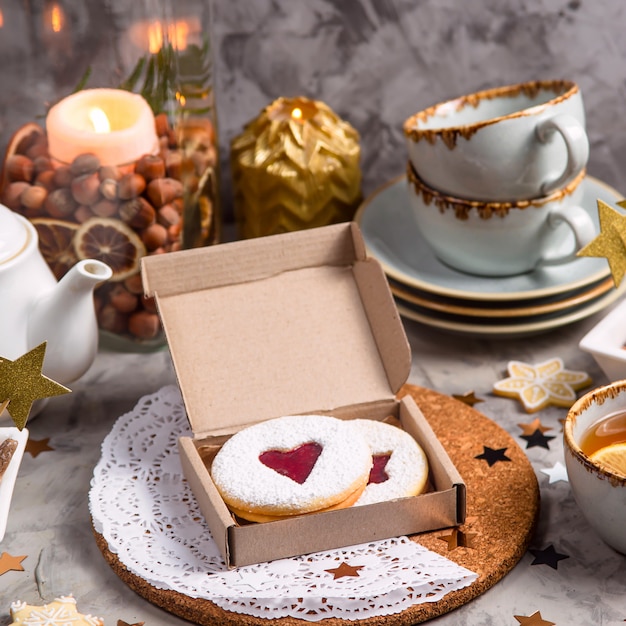 Biscuits ronds avec de la confiture en forme de coeur dans une boîte cadeau parmi des décorations de Noël et des bougies sur une table grise