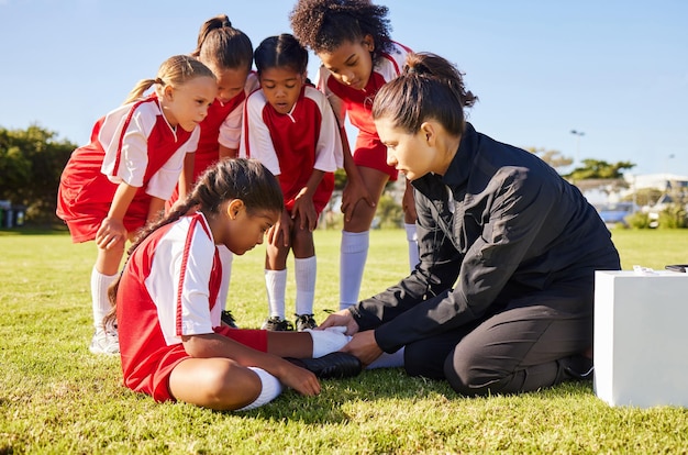 Blessures sportives et équipe de football pour enfants avec leur entraîneur dans un caucus aidant une athlète féminine Entraînement physique et enfant souffrant de douleurs ou d'entorses musculaires après un match sur un terrain de football extérieur