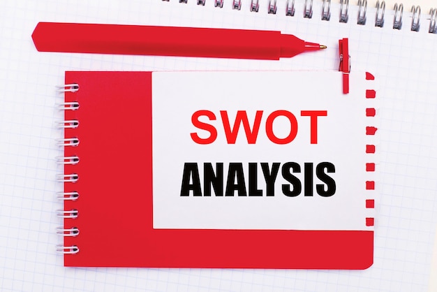 Sur un bloc-notes blanc, un stylo rouge, un bloc-notes rouge et une feuille de papier blanche avec le texte ANALYSE SWOT