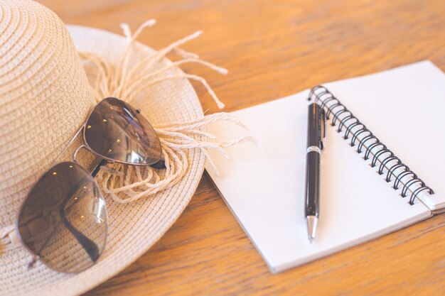 Bloc-notes sur écran blanc avec stylo, chapeau de paille, lunettes de soleil sur le bureau en bois