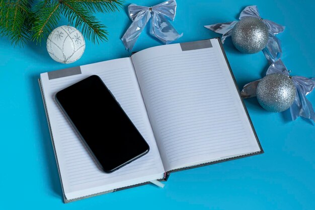 Bloc-notes gris et téléphone noir entourés de décorations de Noël scintillantes argentées sur fond bleu vue de dessus