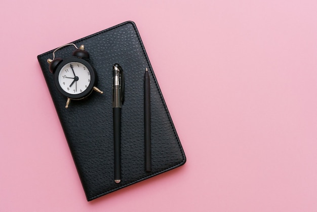 Bloc-notes noir avec un stylo et un réveil sur une surface rose