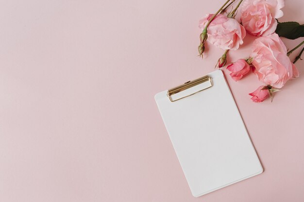 Bloc-notes de presse-papiers de feuille de papier vierge, bouquet de fleurs roses sur fond rose. Flatlay, vue de dessus