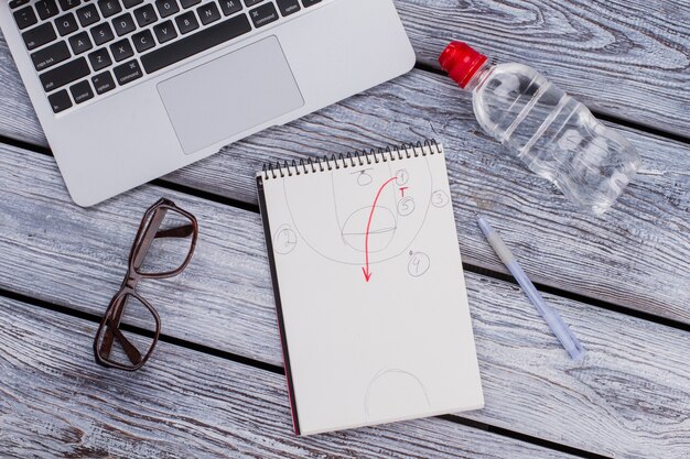 Bloc-notes avec schéma de plan et accessoires de travail de bureau. Vue de dessus à plat. Bouteille en plastique avec de l'eau et un ordinateur portable sur une table en bois blanche.