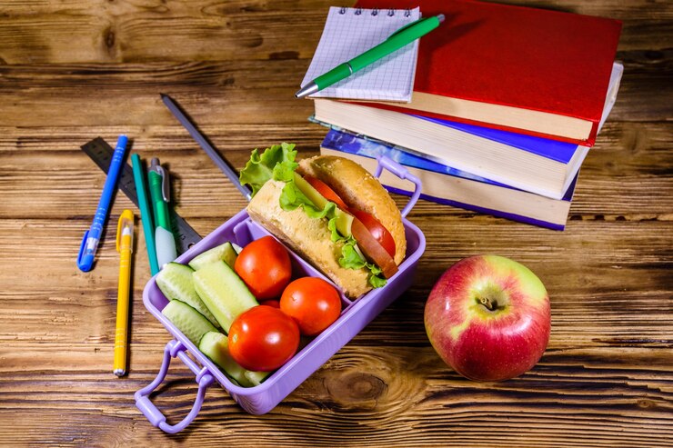 Photo bloc-notes, stylos, pomme mûre, pile de livres et boîte à lunch avec hamburger, concombres et tomates sur une table en bois rustique