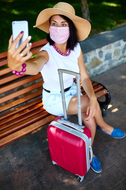 Blogger femme en masque de protection, dans le parc en plein air avec une valise, prenez selfie