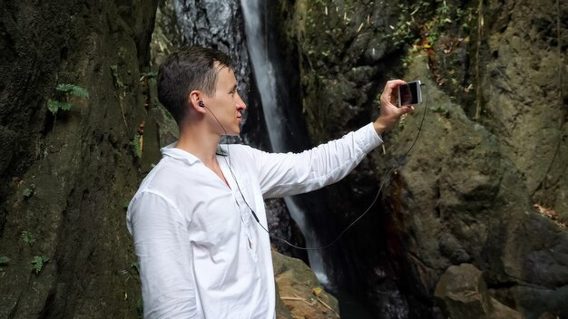 Un blogueur aux cheveux noirs en chemise blanche utilise un casque tenant un smartphone et passe un appel vidéo contre une cascade tropicale dans une réserve vue rapprochée
