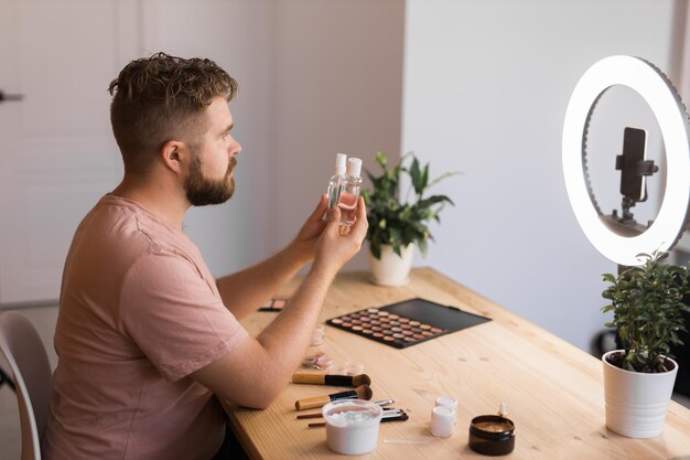 Blogueur de beauté masculine avec produit de maquillage enregistrant une vidéo à la maison à l'aide d'une lampe annulaire et d'un vlog pour smartphone