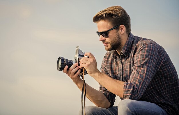 Blogueur tirant vlog Beau blogueur mec voyageur appareil photo rétro Réglages manuels Concept de photojournaliste Blogueur de voyage Reporter prenant une photo Guy à l'extérieur fond de ciel bleu Équipement vintage