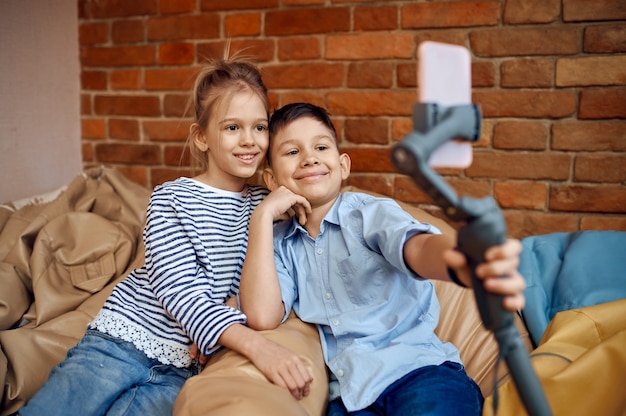 Des blogueurs d'enfants souriants font des selfies sur un canapé, de petits vloggers. Enfants bloguant en home studio, médias sociaux pour jeune public, diffusion Internet en ligne