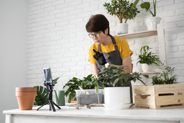 Une blogueuse assise devant la caméra d'un smartphone sur un trépied enregistre une vidéo didactique pédagogique pour son blog tourne le processus de replantation de fleurs et de plantes vertes pleines de terre aime le passe-temps botanique