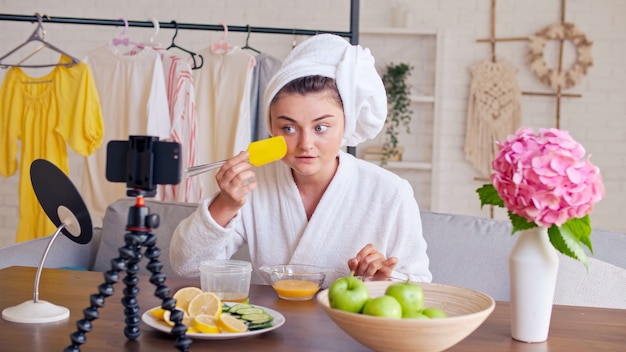 Une blogueuse beauté vêtue d'une robe blanche avec une serviette blanche sur la tête applique un masque hydratant vert sur...