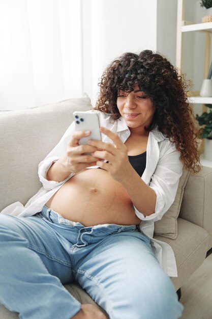 Une blogueuse enceinte est assise sur le canapé à la maison et prend des photos d'elle-même sur le téléphone.