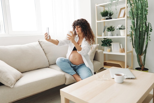 Une blogueuse enceinte avec un pot de pilules et de vitamines est assise sur le canapé à la maison et prend des photos.