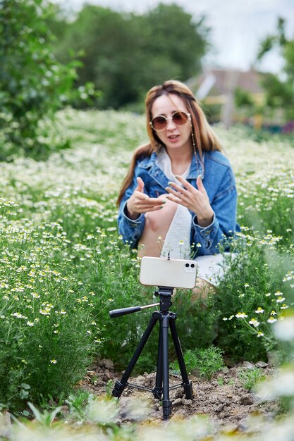 Une blogueuse franche et insouciante du millénaire qui enregistre une vidéo à l'aide d'un trépied et d'un téléphone avec appareil photo dans des fleurs de camomille Mode de vie des blogs Utilisation de la technologie moderne