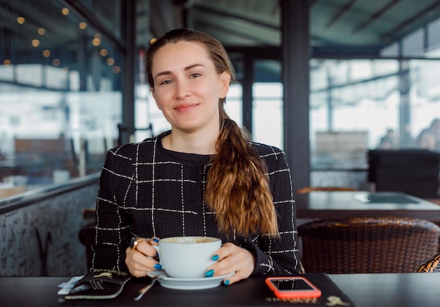 Une blogueuse souriante regarde la caméra en tenant une tasse en s'asseyant dans un café