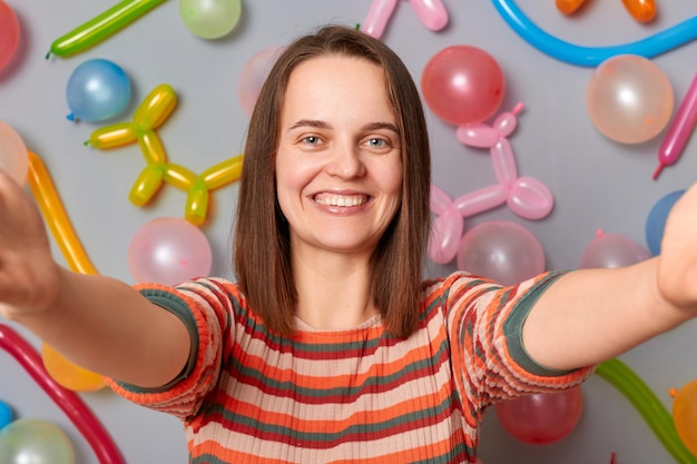 Une blogueuse souriante vêtue d'une robe rayée debout contre un mur gris avec une décoration de ballons faisant un selfie POV diffusant en direct de sa fête d'anniversaire