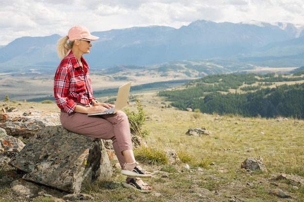 Une blogueuse voyageuse mature travaille à distance sur un ordinateur netbook tout en profitant de la vue sur le paysage de la nature des montagnes à l'extérieur