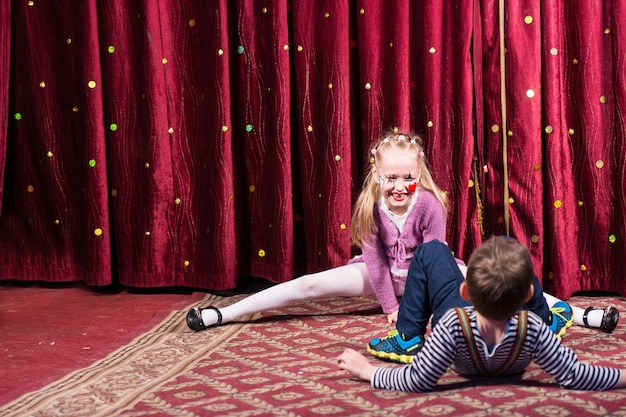 Blond Girl Wearing Clown Make Up Performing Splits sur scène avec rideau rouge avec jeune garçon en premier plan