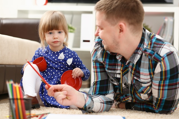 Blond petite fille souriante tenir au crayon de bras dessiner quelque chose avec papa. Belle femme jeune artiste mineur baby-sitter art joie développement juvénile parent style de vie jeune peintre