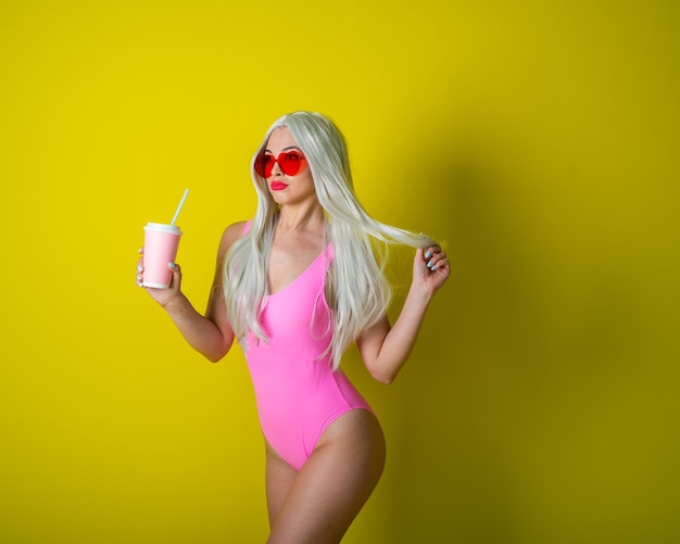 La blonde avec un beau gros cul en maillot de bain rose boit un verre Fille en perruque posant sur fond jaune Une femme aux longs cheveux artificiels en lunettes de soleil à la vue des coeurs