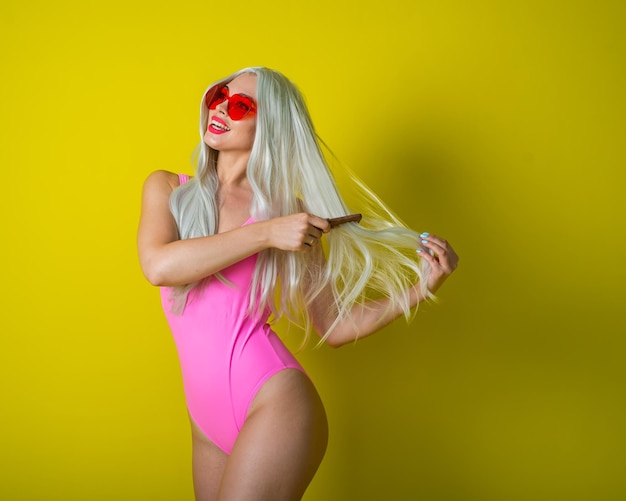 La blonde avec une belle silhouette en maillot de bain rose se peigne Fille dans une perruque posant sur un fond jaune Une femme aux longs cheveux artificiels tient un peigne