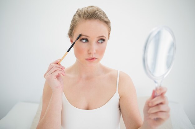 Blonde calme naturelle tenant le miroir et à l&#39;aide d&#39;une brosse à sourcils