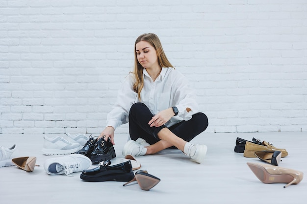 Blonde élégante choisissant des chaussures assises sur le sol Vue latérale d'un grand nombre de chaussures pour femmes sur le sol un choix de chaussures élégantes