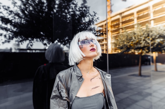 Blonde élégante dans des vêtements futuristes et des lunettes sur un miroir.