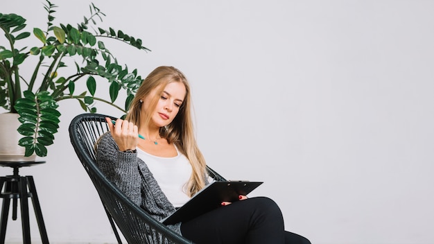 Photo blonde jeune femme assise sur une chaise écrivant des notes sur le presse-papiers contre le mur blanc