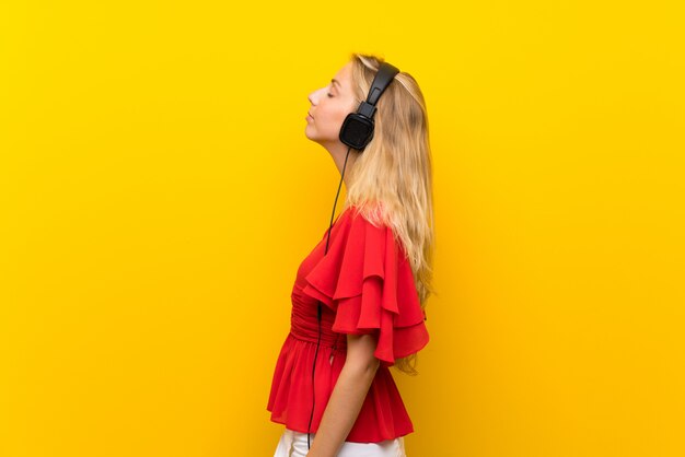 Blonde jeune femme sur un mur jaune, écouter de la musique avec des écouteurs