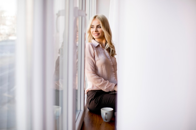 Blonde jeune femme se détendre et boire un café par la fenêtre