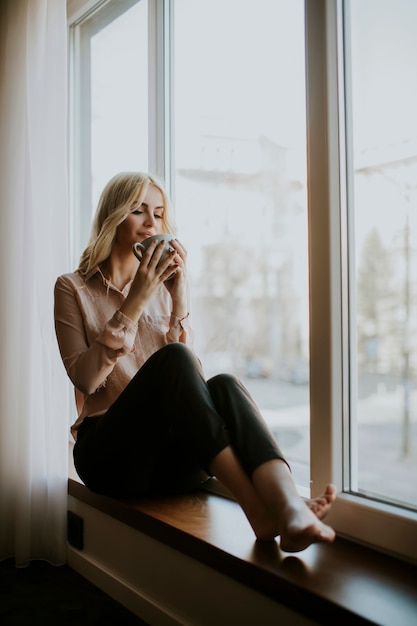 Blonde jeune femme se détendre et boire du café par la fenêtre