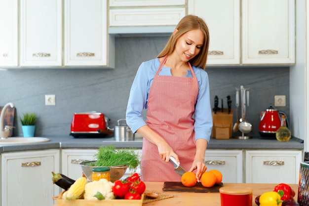Blonde jeune femme en tablier coupe des oranges dans sa cuisine