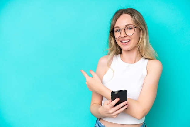 Blonde jeune fille anglaise isolée sur fond bleu à l'aide d'un téléphone portable et pointant vers l'arrière