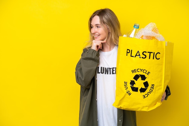 Blonde jeune fille anglaise tenant un sac plein de bouteilles en plastique à recycler isolé sur fond jaune en pensant à une idée et en regardant de côté