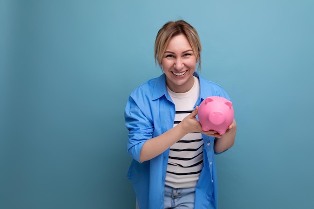 Blonde jolie jeune femme en tenue décontractée avec un sourire tenant une tirelire avec des économies en elle