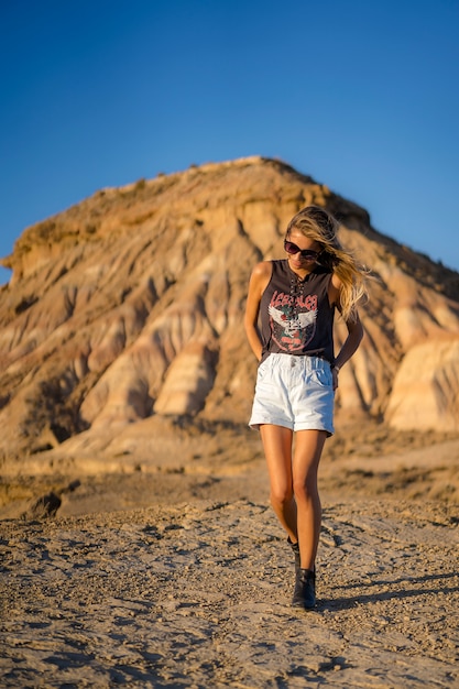 Blonde avec des lunettes de soleil, des bottes noires et un short se promenant dans le désert