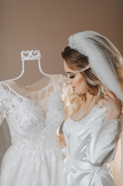 La blonde mariée porte une robe de satin, de beaux cheveux et un maquillage, un buste ouvert, un portrait de mariage, un sourire sincère, une belle mariée en robe de chambre pose à côté de sa robe.