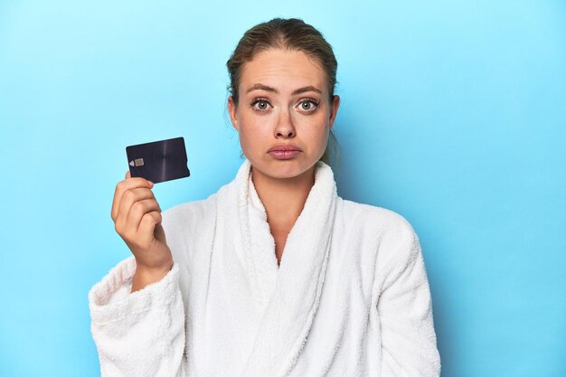 Blonde en peignoir avec carte de crédit en studio bleu hausse les épaules et ouvre les yeux confus