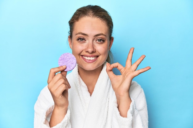 Blonde en peignoir avec éponge faciale en studio joyeuse et confiante montrant un geste correct